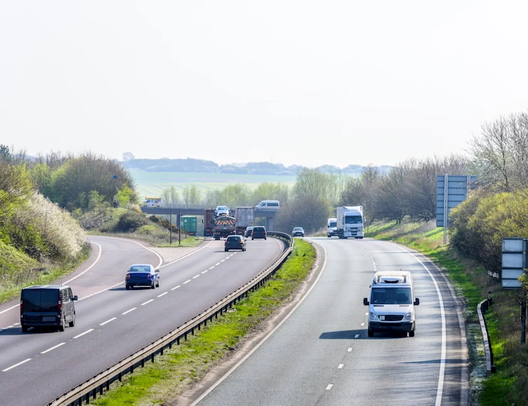 A busy motorway with cars, trucks, and vans driving on it with green location pinpoint icons above three vehicles.